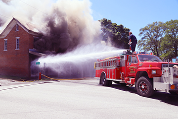 Turning up the volume — Arenzville Firefighter Lee Burrus mans the water cannon atop a pumper truck and pours high volume water into the front of Reg’s Auto Service in an attempt to knock down the flames after fire erupted in the building late Friday morning. Firefighters from Arenzville, Virginia, Meredosia and Beardstown battled the fire in near 90-degree heat. For more photos, please visit our website beardstownnewspapers.com (Photo by Brian DeLoche.)