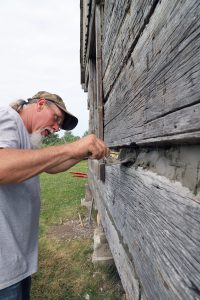 Artist at work — With a pallet of cement and mortar, Ruppert Parrish artfully fills in the gaps between the logs of a cabin wall at The Rexroat Prairie in Virginia. The material between the logs is called chinking, and works to keep wind and moisture out of the cabin and helps prevent decay. At right is the “Dog Trot” cabin, so named because the center space allowed dogs to trot in and out. (Photo by Brian DeLoche.)