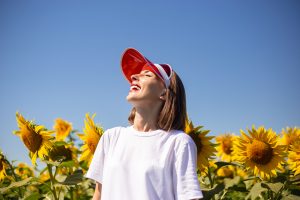 Young woman with closed eyes in red sun visor and white t-shirt looks at the sky on a sunflower field.